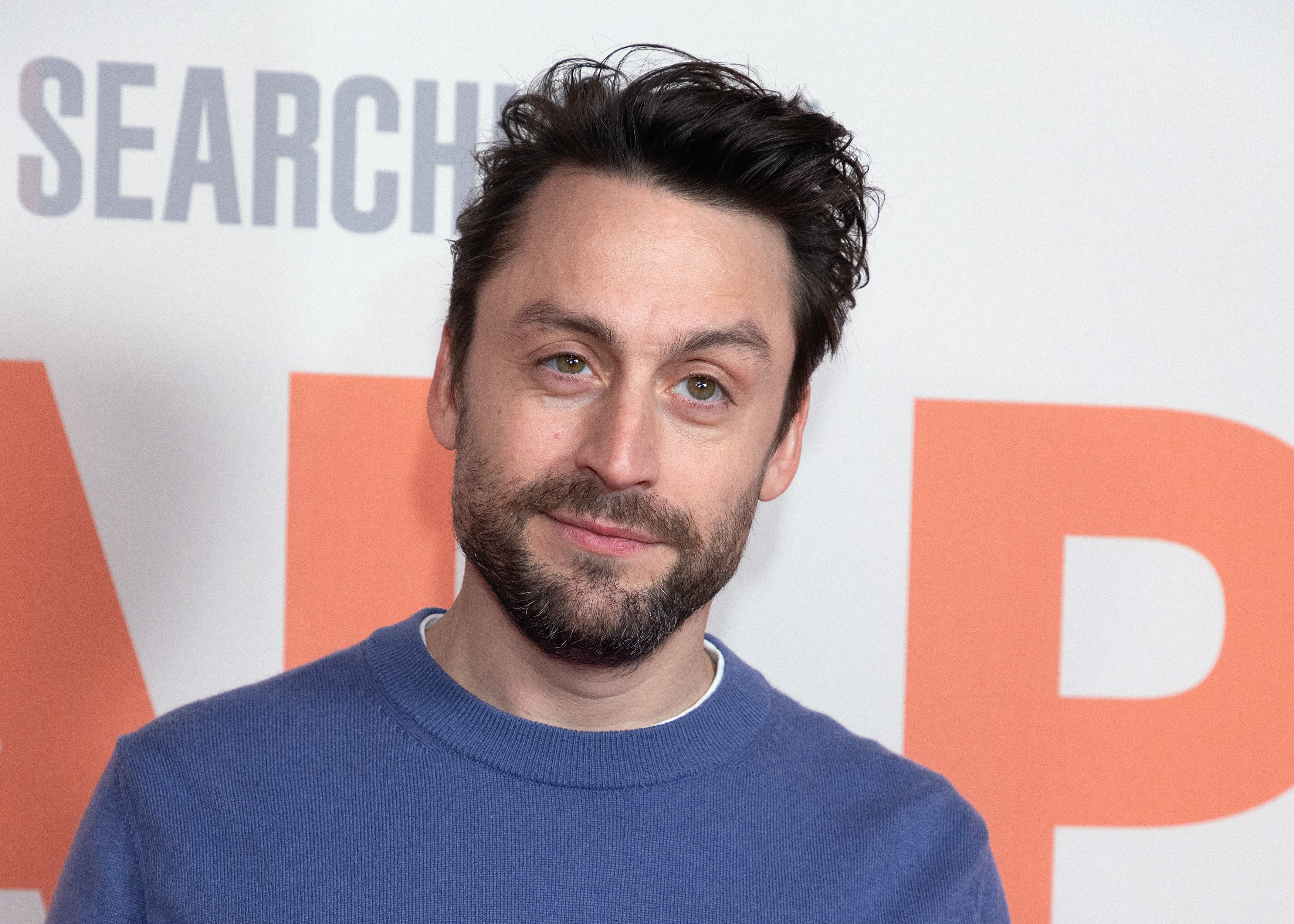 Kieran Culkin with short dark hair and a beard, wearing a blue sweater, stands in front of a backdrop with text.