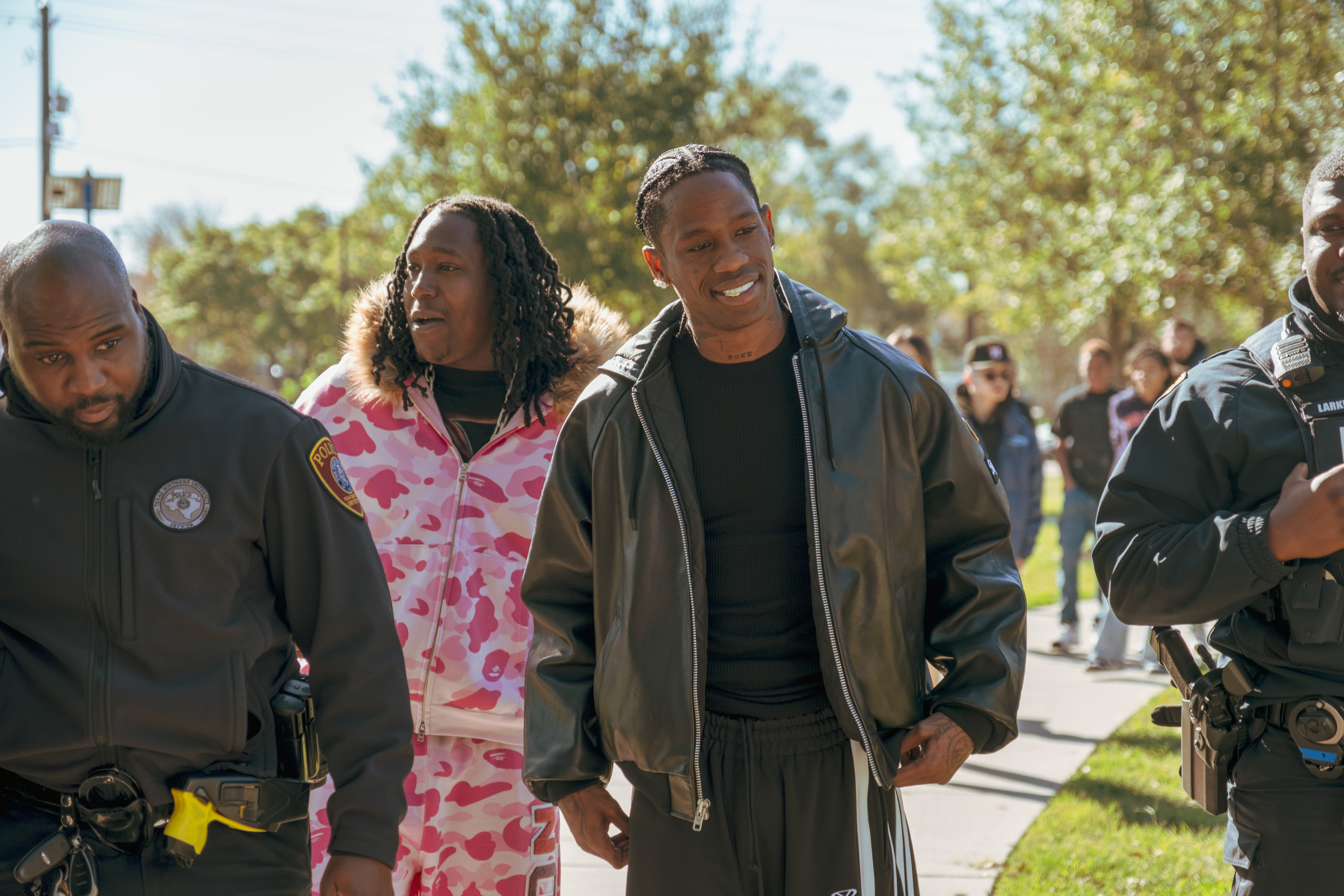 Travis Scott in a black jacket walking outdoors with a group, including a person in a pink camo outfit and police officers.