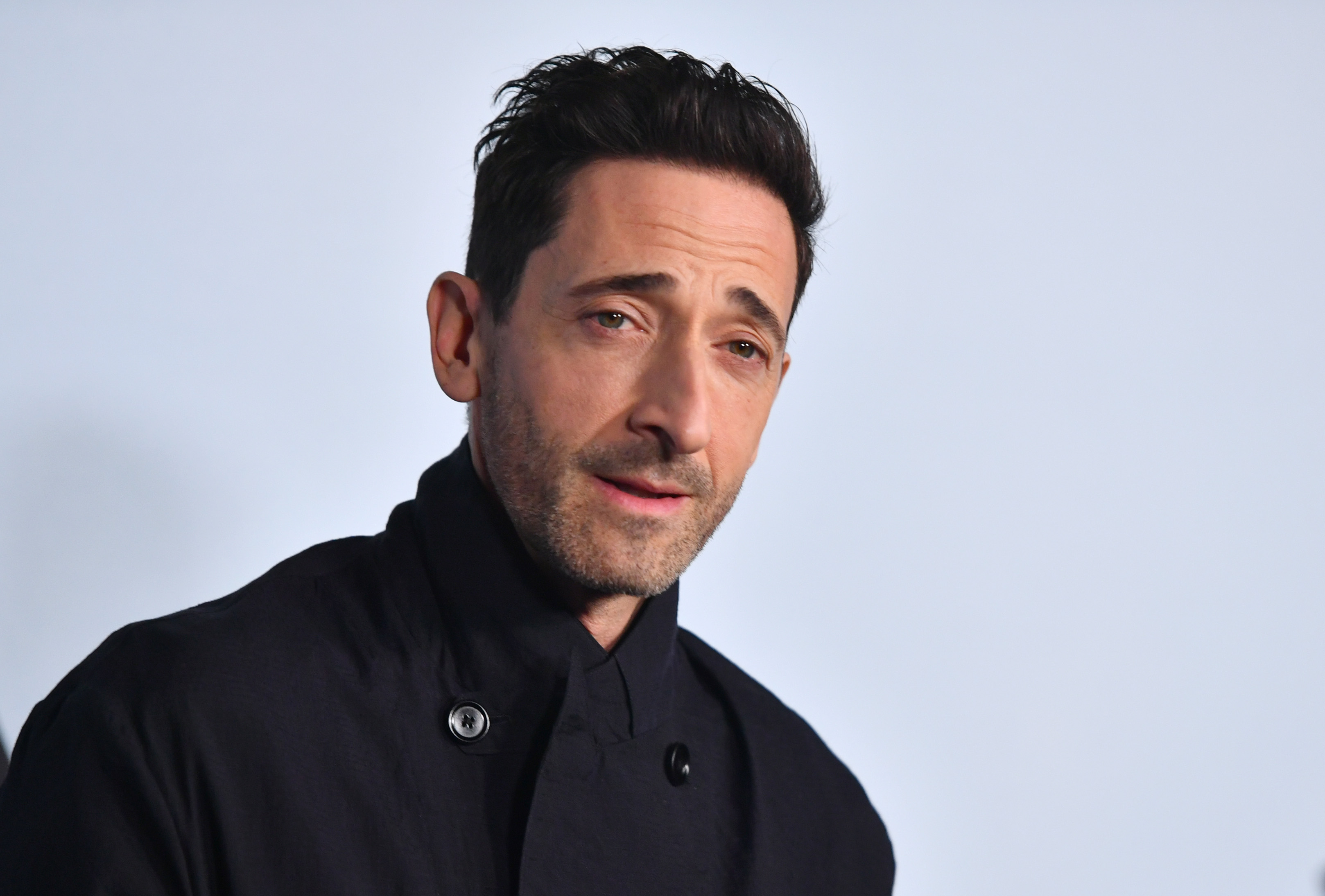 Adrien Brody in a dark coat, looking slightly to the side against a light background.