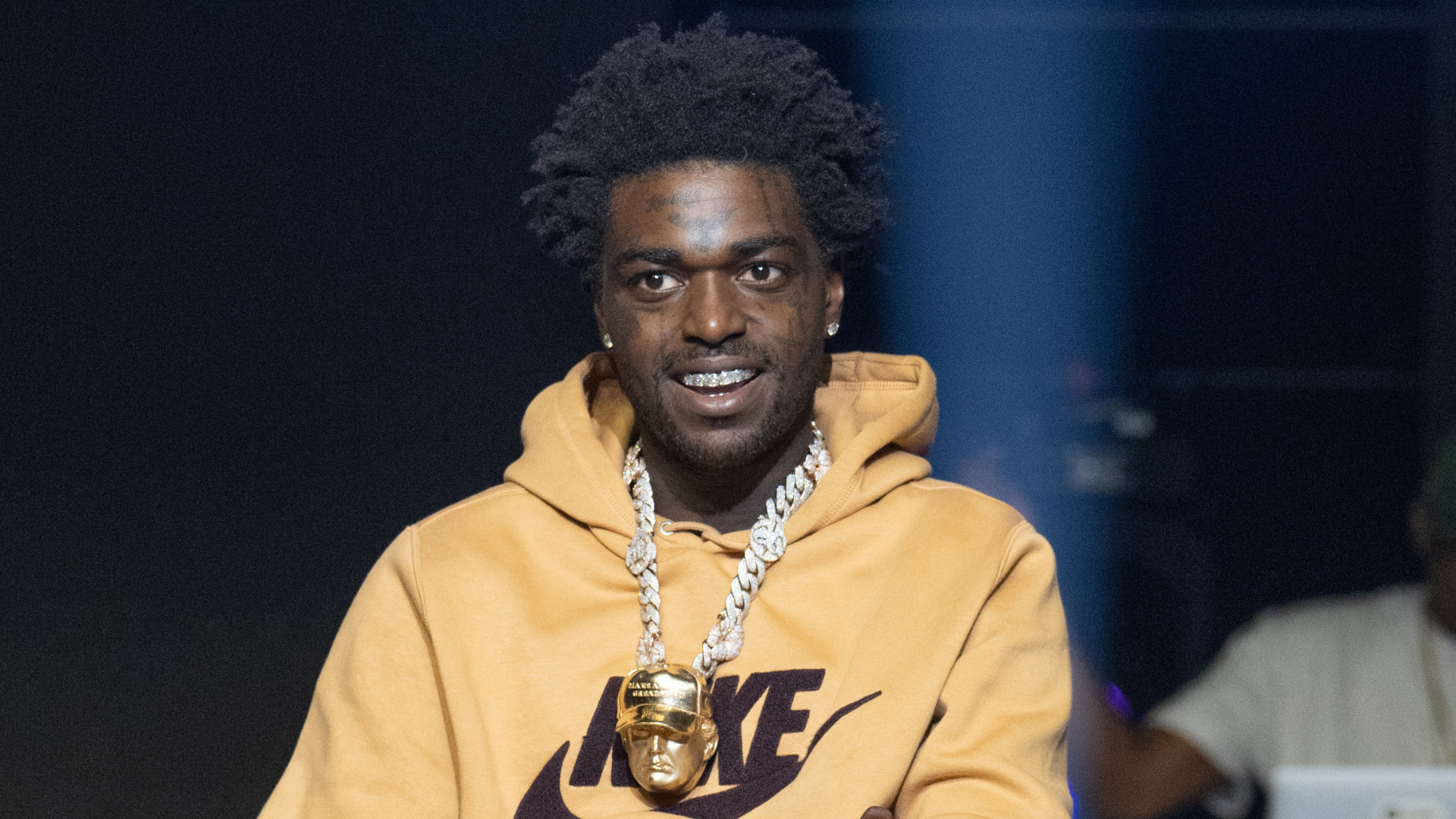 A man with a gold chain and a Nike hoodie smiles, standing under a spotlight.