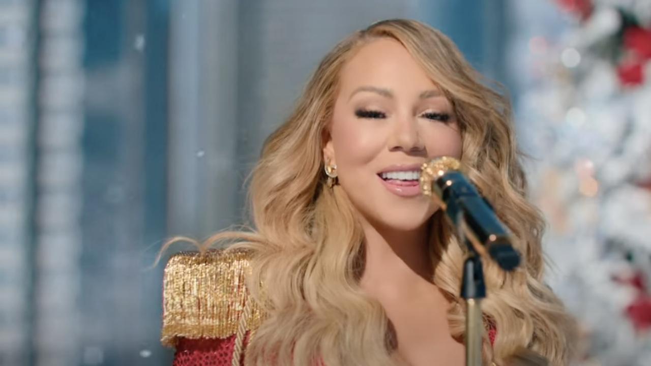 Mariah Carey singing into a microphone, wearing a red and gold outfit with long, wavy blonde hair.