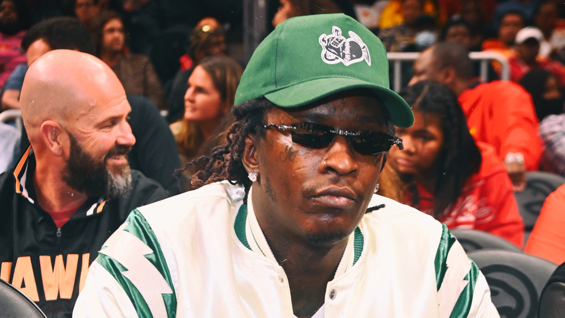 Young Thug wearing a green cap and sunglasses, sitting in a crowd.