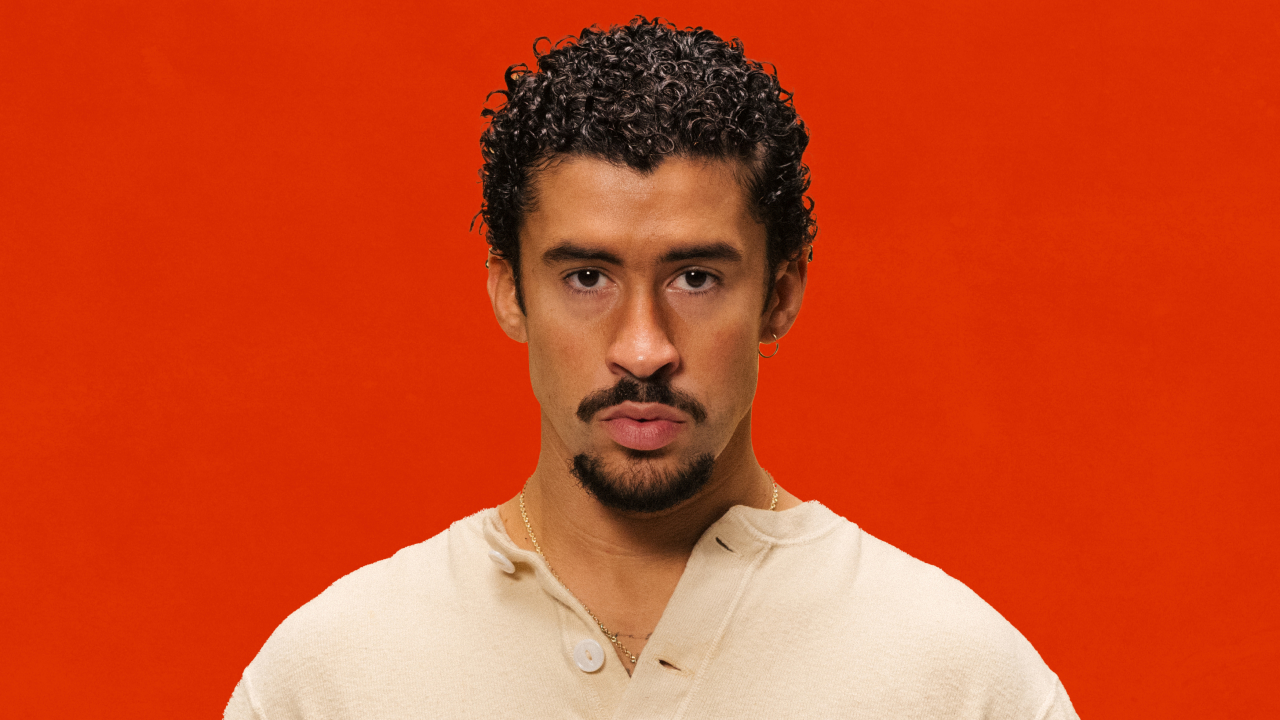 Bad Bunny with curly hair and a goatee, wearing a beige shirt, against an orange background.