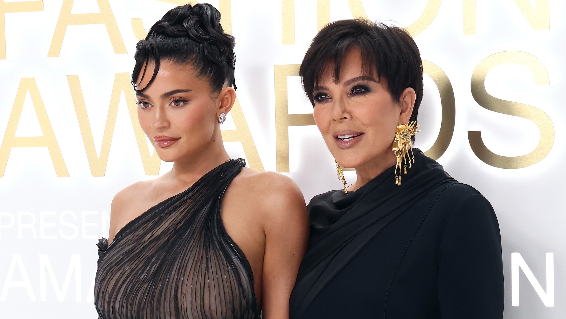 Kylie Jenner and Kris Jenner at a fashion awards event. Kylie wears a sheer black dress, and Kris is in a black outfit with gold earrings.