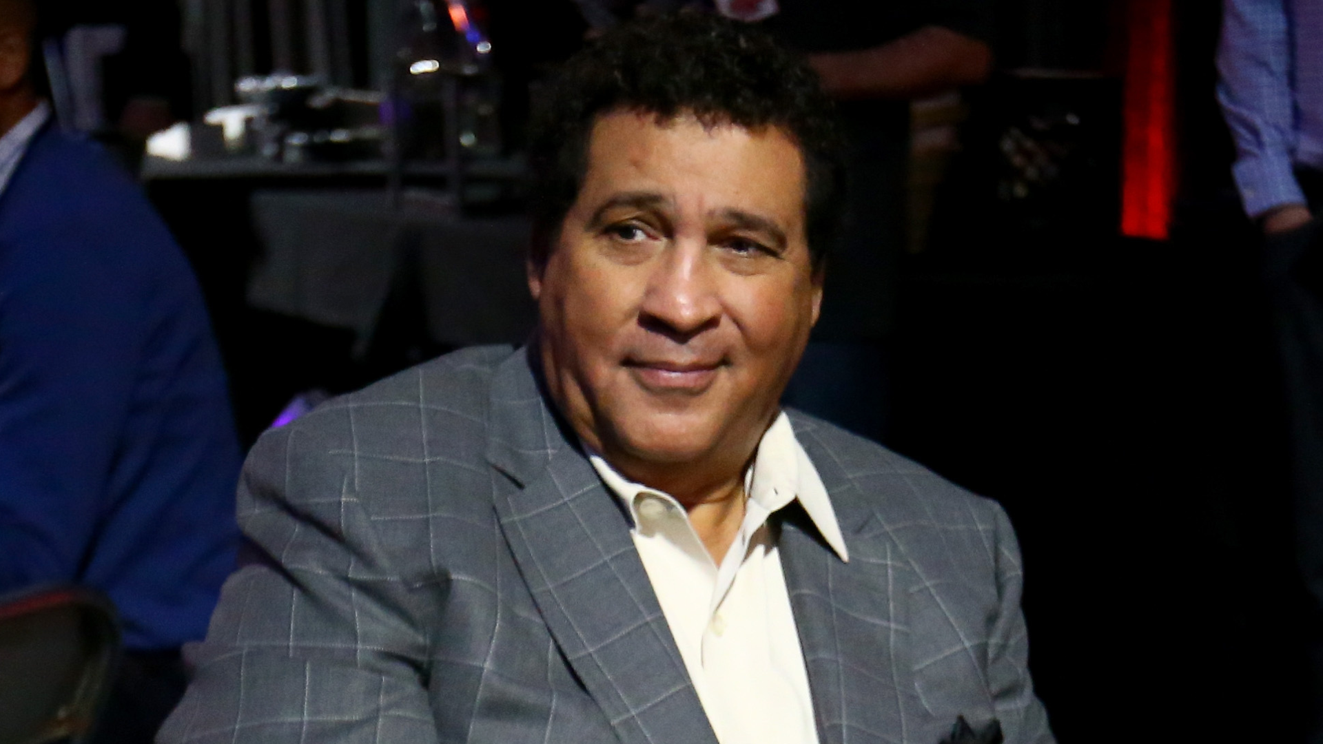 A man in a gray plaid suit sits at an event, looking to the side with a neutral expression.