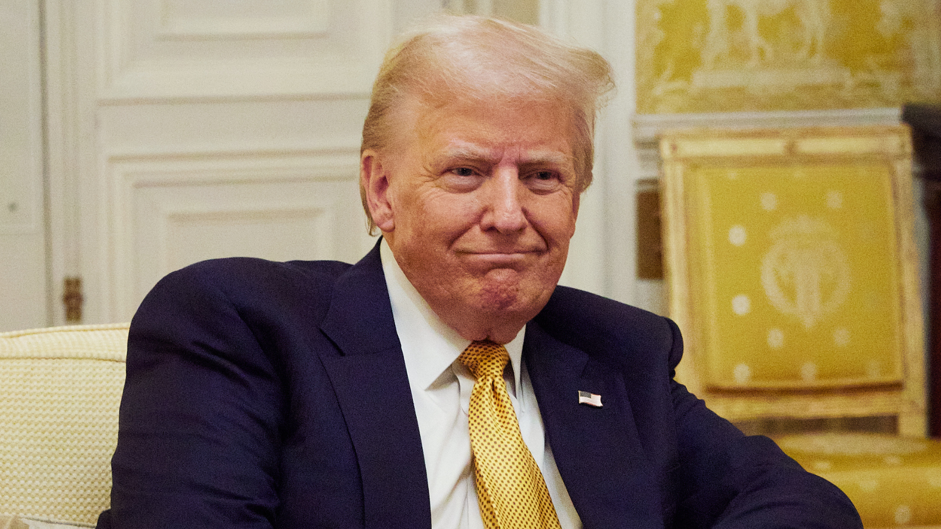 Donald Trump sitting in a room with ornate decor, wearing a dark suit and yellow tie, looking slightly to the side.