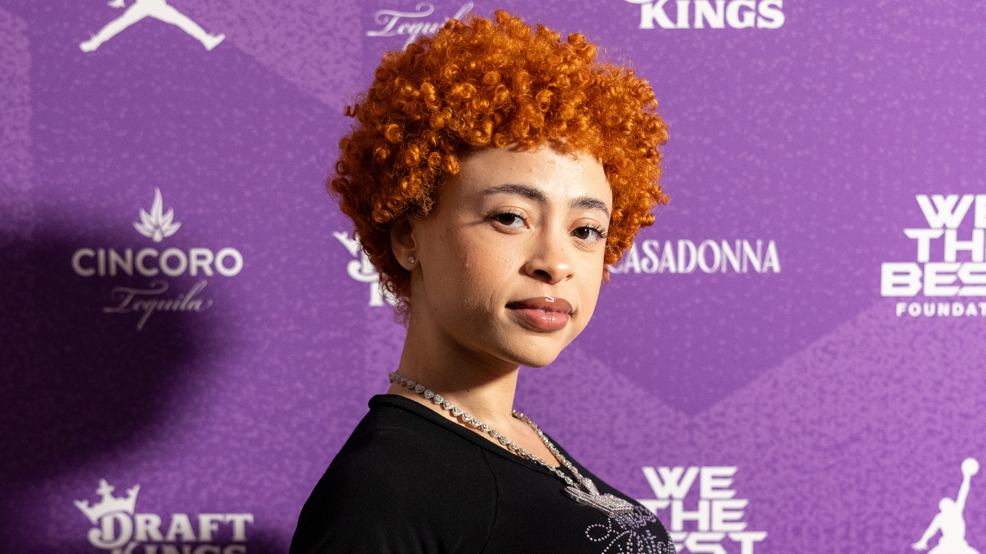 A woman with curly orange hair poses against a purple backdrop with logos and text.