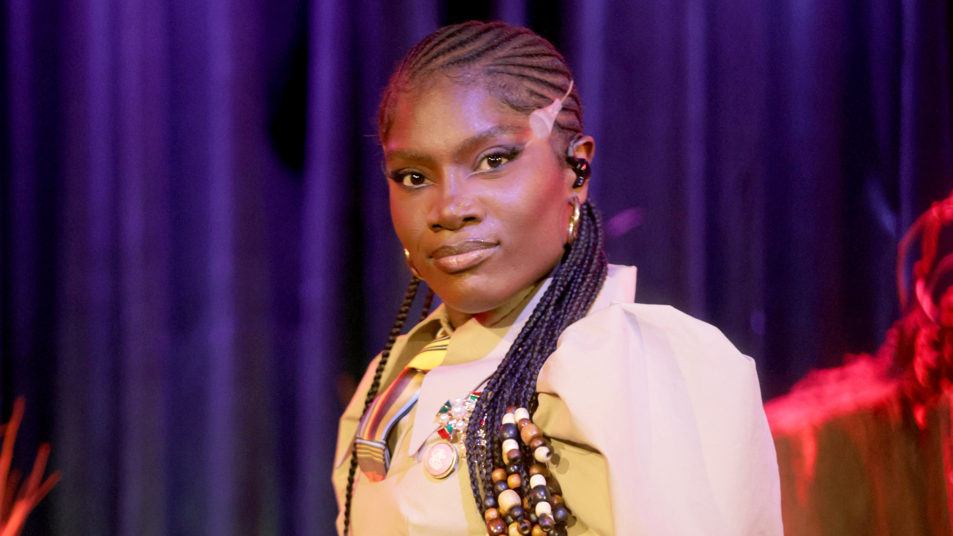 A person with braided hair and colorful beads, wearing a light-colored outfit with a tie, stands in front of a purple curtain.