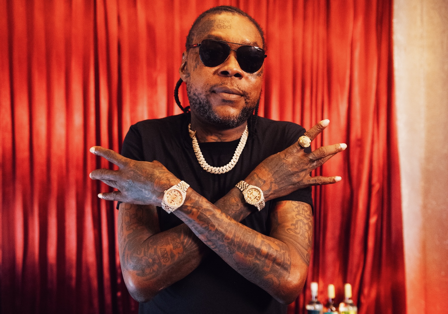Vybz Kartel with tattoos, wearing sunglasses, a black shirt, and jewelry, poses with crossed arms in front of red curtains.