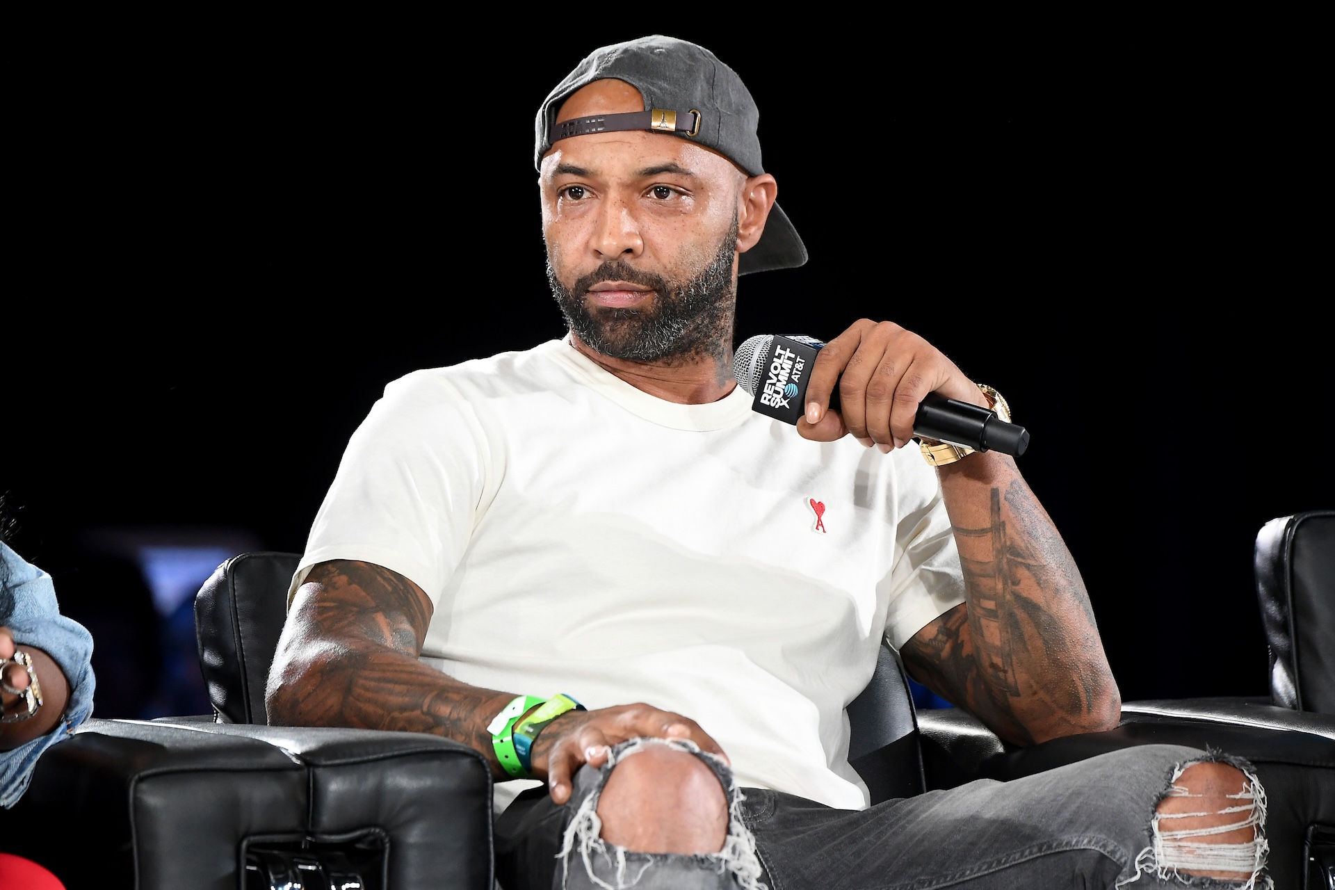 Joe Budden with a beard and tattoos, wearing a cap backwards, holds a microphone while sitting on a stage in a casual outfit.