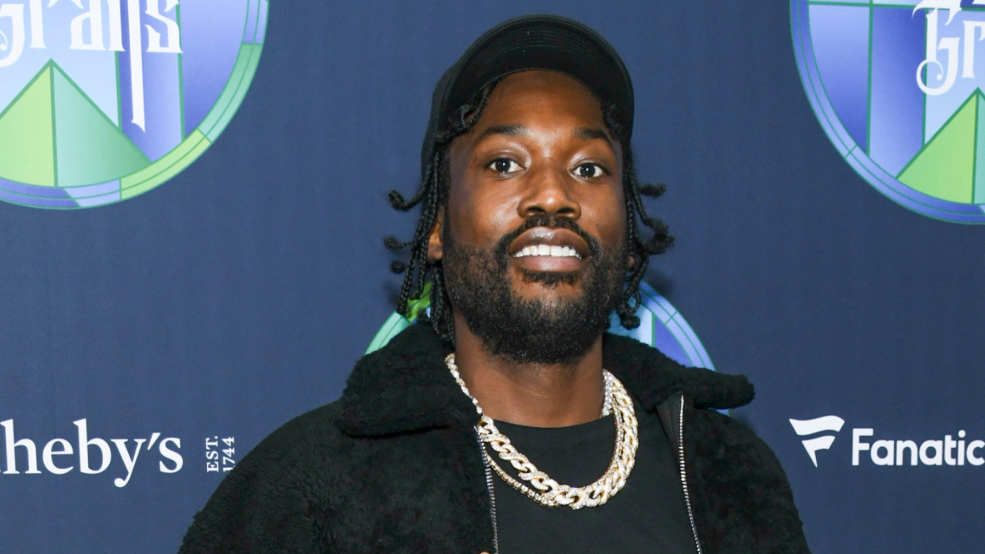 Meek Mill, wearing a black jacket and gold chain, smiles at an event with branded backdrops.