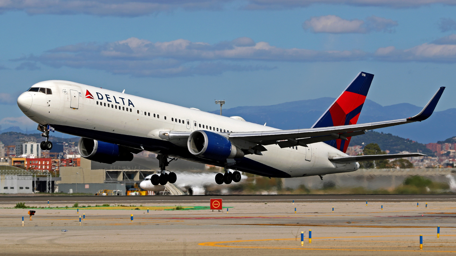 Delta Airlines plane taking off, with mountains and a cityscape in the background.
