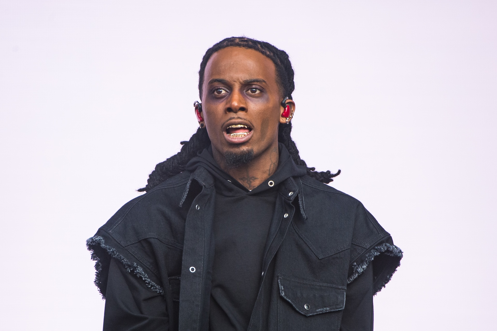 Playboi Carti with long hair and gold teeth, wearing a black jacket and hoodie, stands against a plain background.