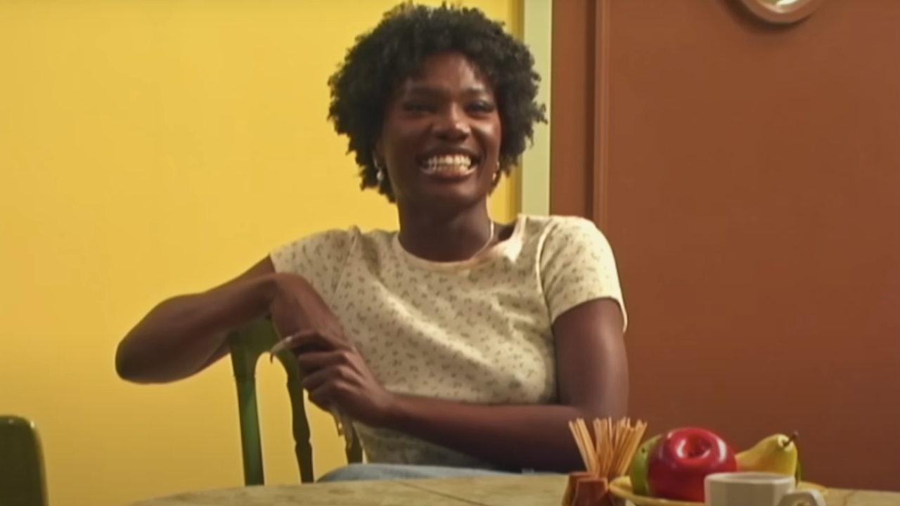 Doechii sits at a table, smiling. The room has a yellow wall, and there's a fruit bowl and cup on the table.