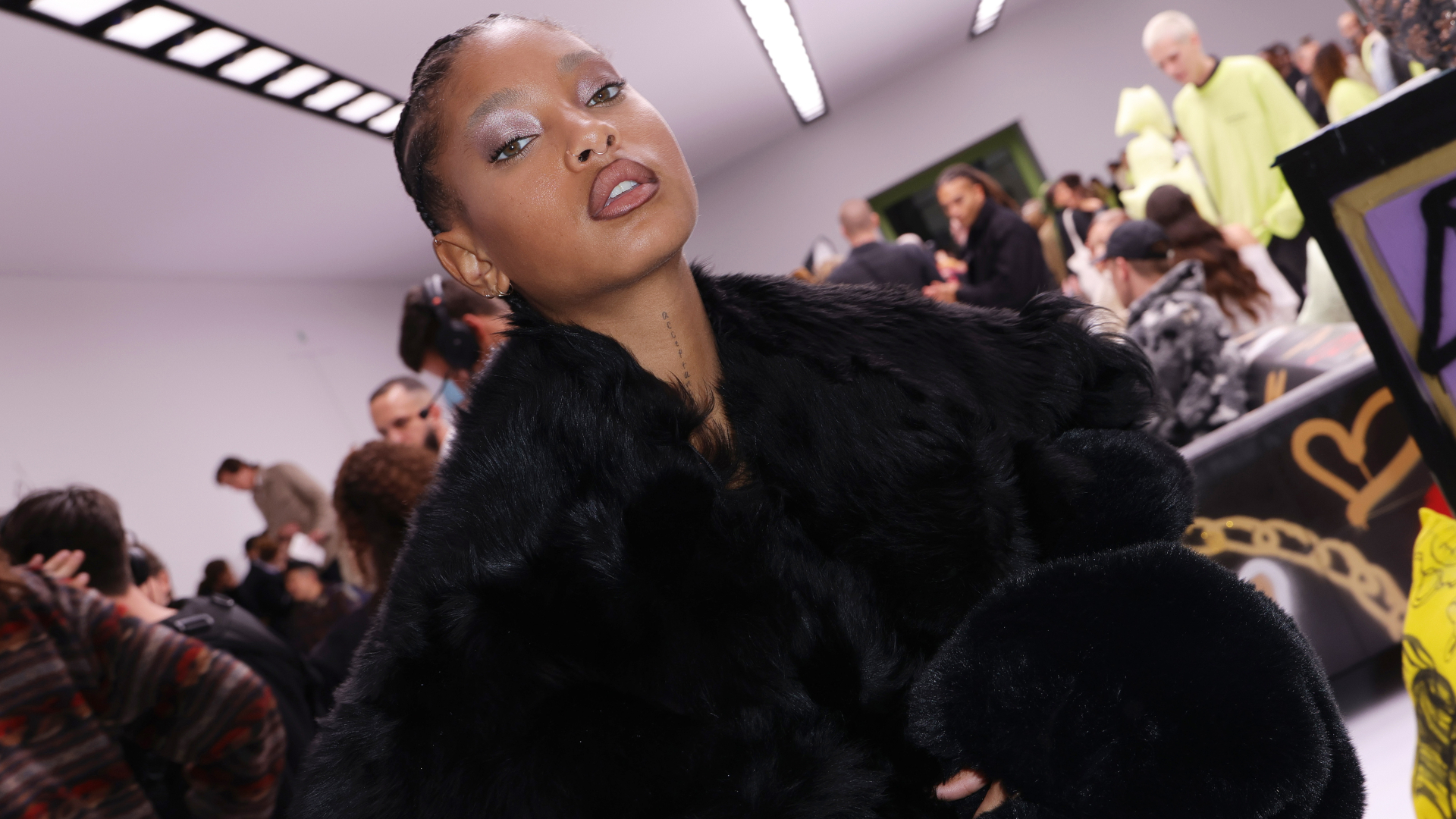 A woman in a black fur coat poses confidently in a crowded room with people in the background.
