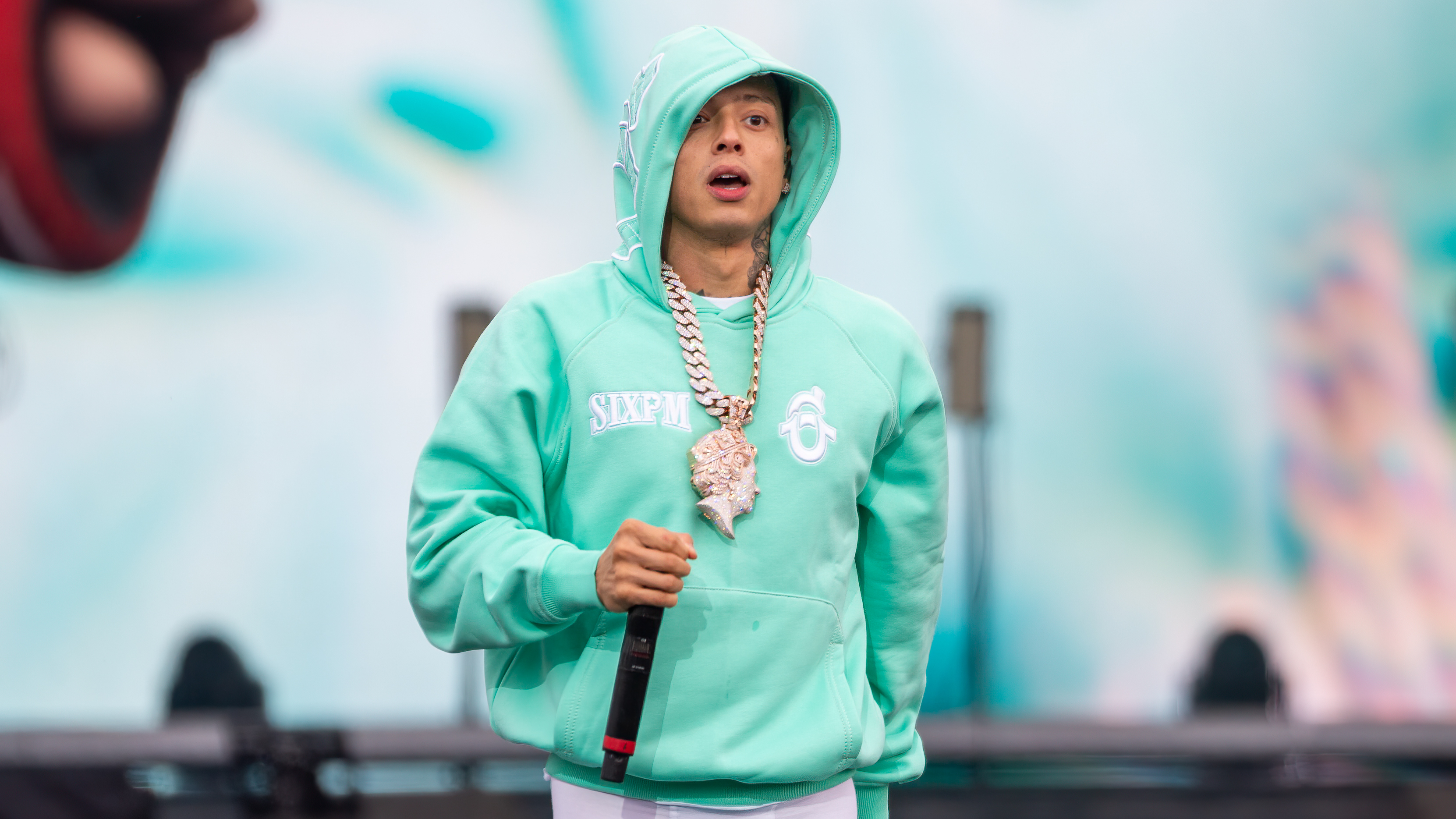 A person in a mint green hoodie with a large chain necklace holds a microphone, performing on stage with a blurred background.
