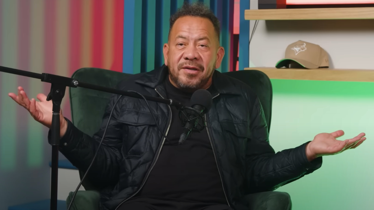 A man is sitting in a chair with his arms raised, speaking into a microphone. The background has colorful vertical stripes.