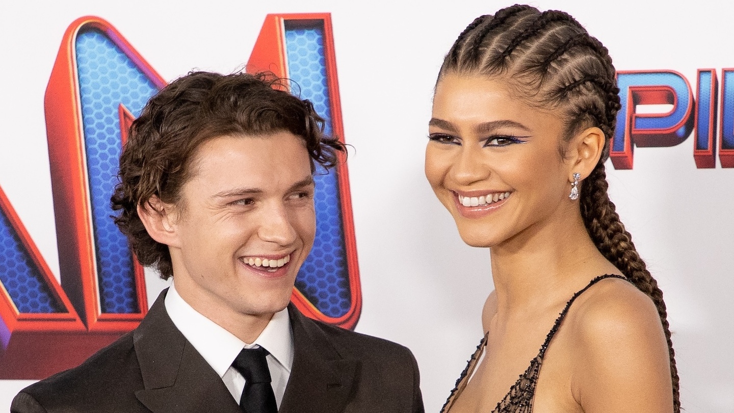 Tom Holland and Zendaya at a movie premiere, smiling together. Tom is in a suit, and Zendaya wears a dress with braided hair.