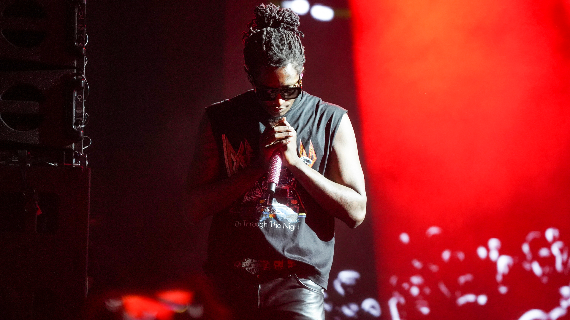 Young Thug wearing sunglasses and a sleeveless shirt is holding a microphone on stage with red lighting in the background.