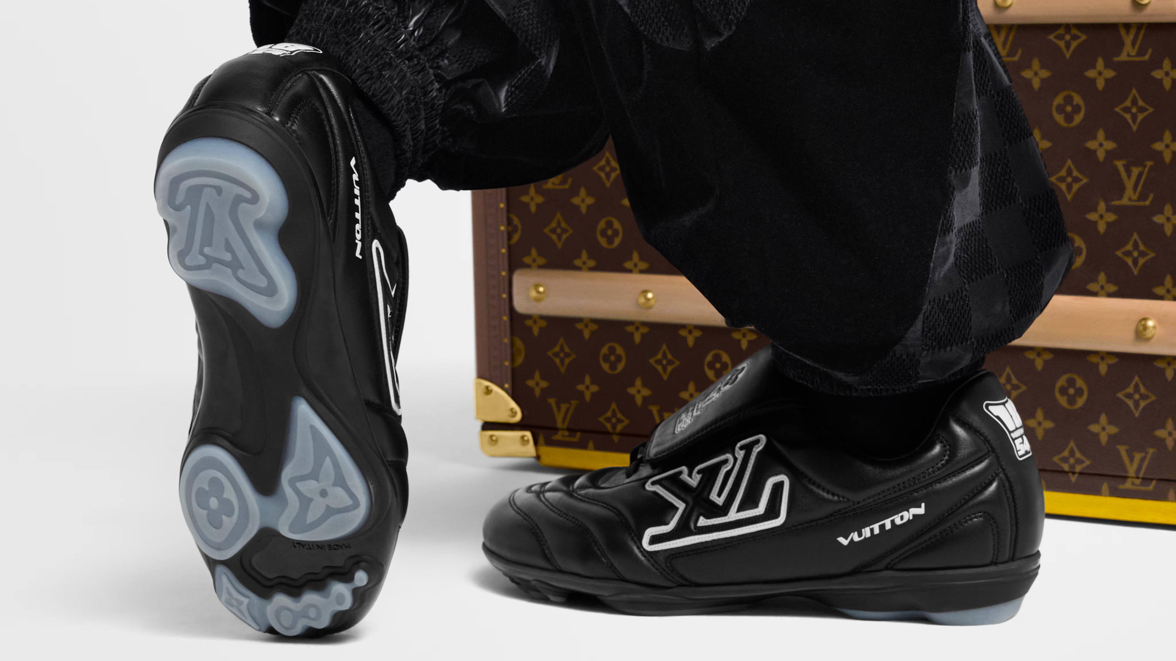 Black Louis Vuitton sneakers with LV logos, worn by a person in black pants, next to a Louis Vuitton monogrammed trunk.