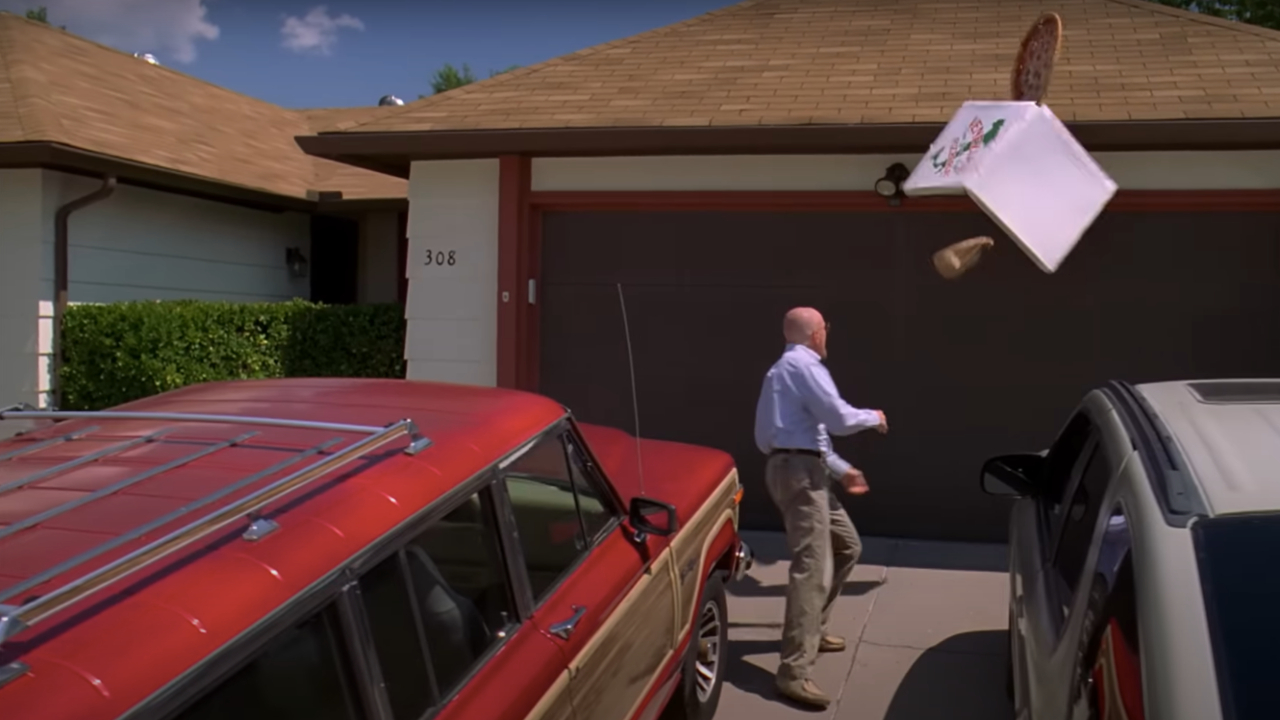 Walter White throws a pizza onto the roof of a suburban house, with two cars parked in the driveway.