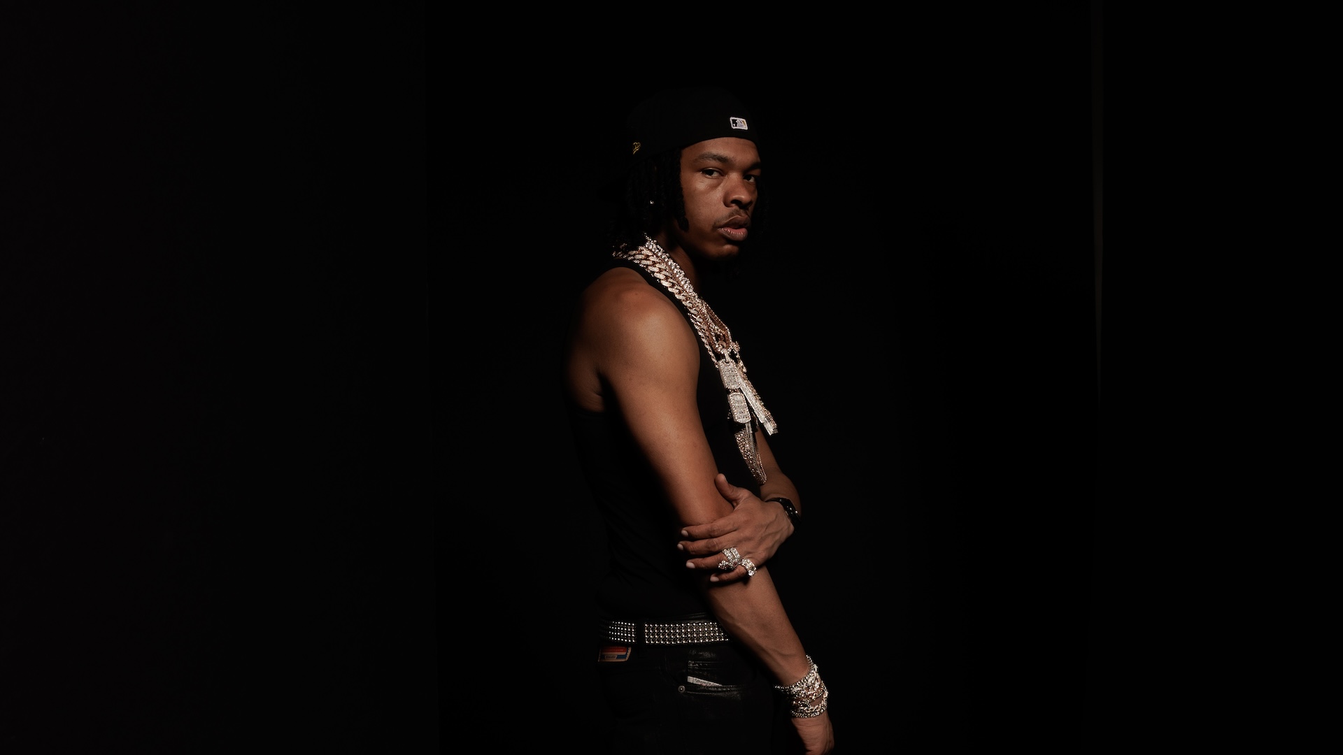 A man in a black tank top and cap stands against a dark background, wearing multiple chains and bracelets, looking to the side.