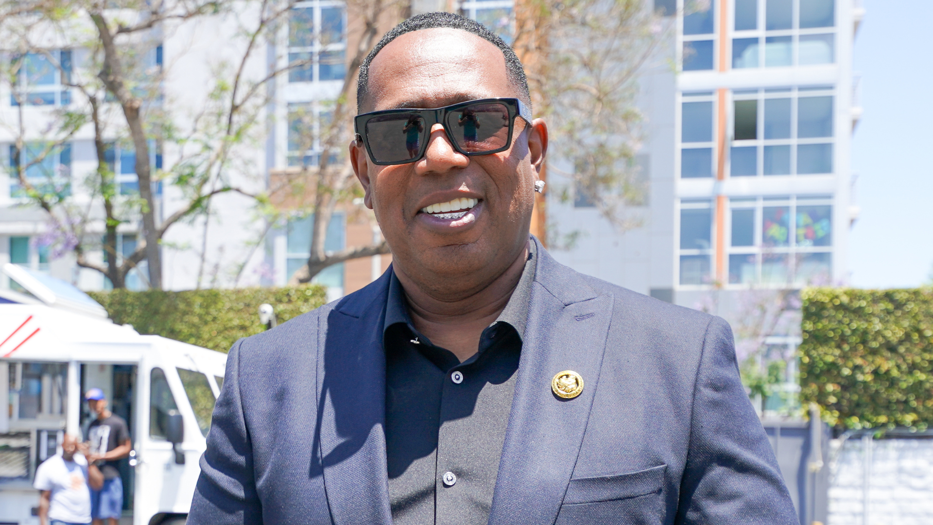 Man wearing sunglasses and a suit, standing outdoors with buildings and trees in the background.