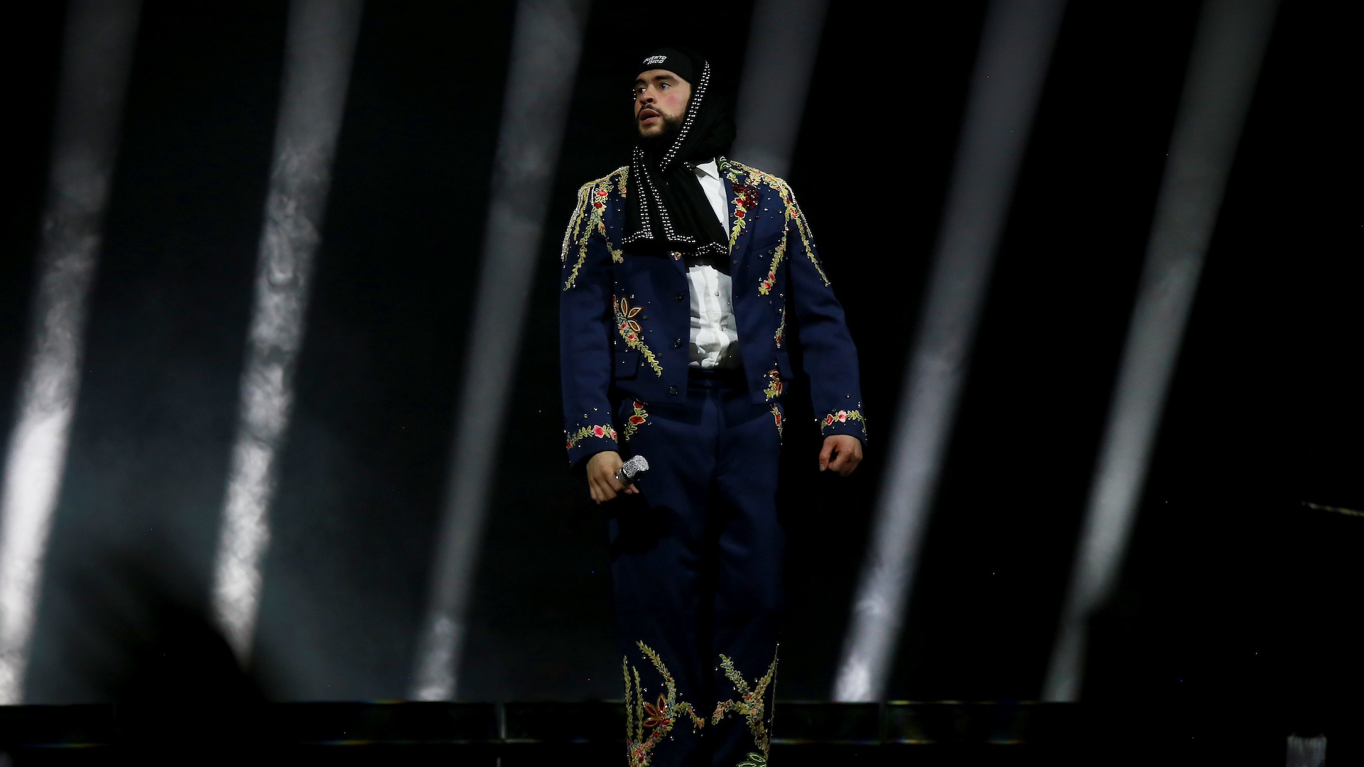 Bad Bunny in a dark, embroidered outfit and headscarf stands on stage, holding a microphone, with dramatic lighting in the background.