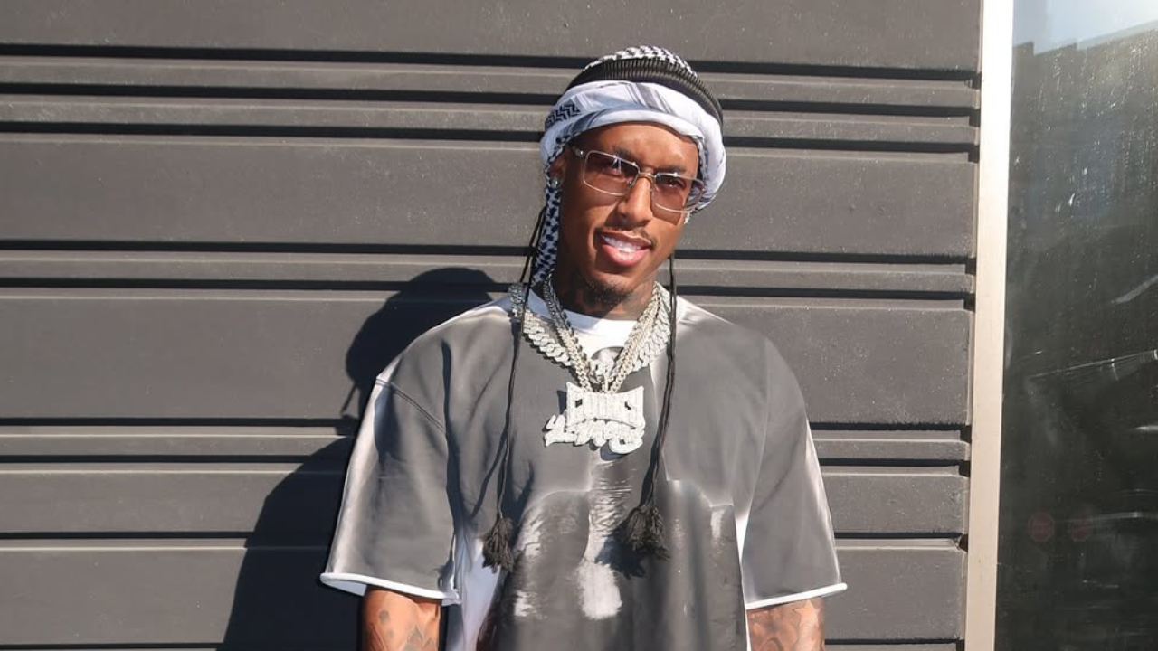 A man wearing sunglasses, a headscarf, and a graphic T-shirt stands against a dark wall, smiling.