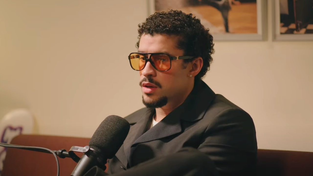 Bad Bunny with curly hair and sunglasses speaks into a microphone, wearing a dark blazer.