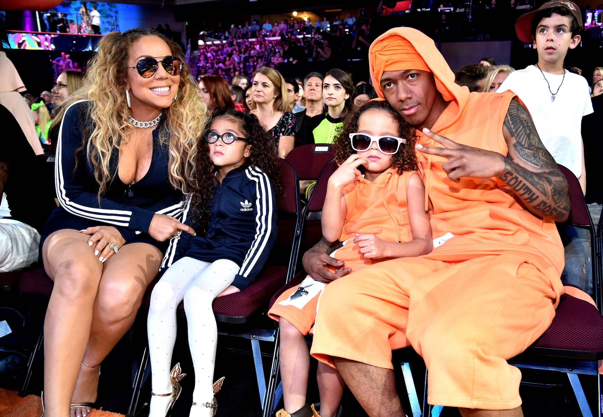 Mariah Carey and Nick Cannon sit with their children at an event. Mariah is in a black outfit, and Nick wears orange.