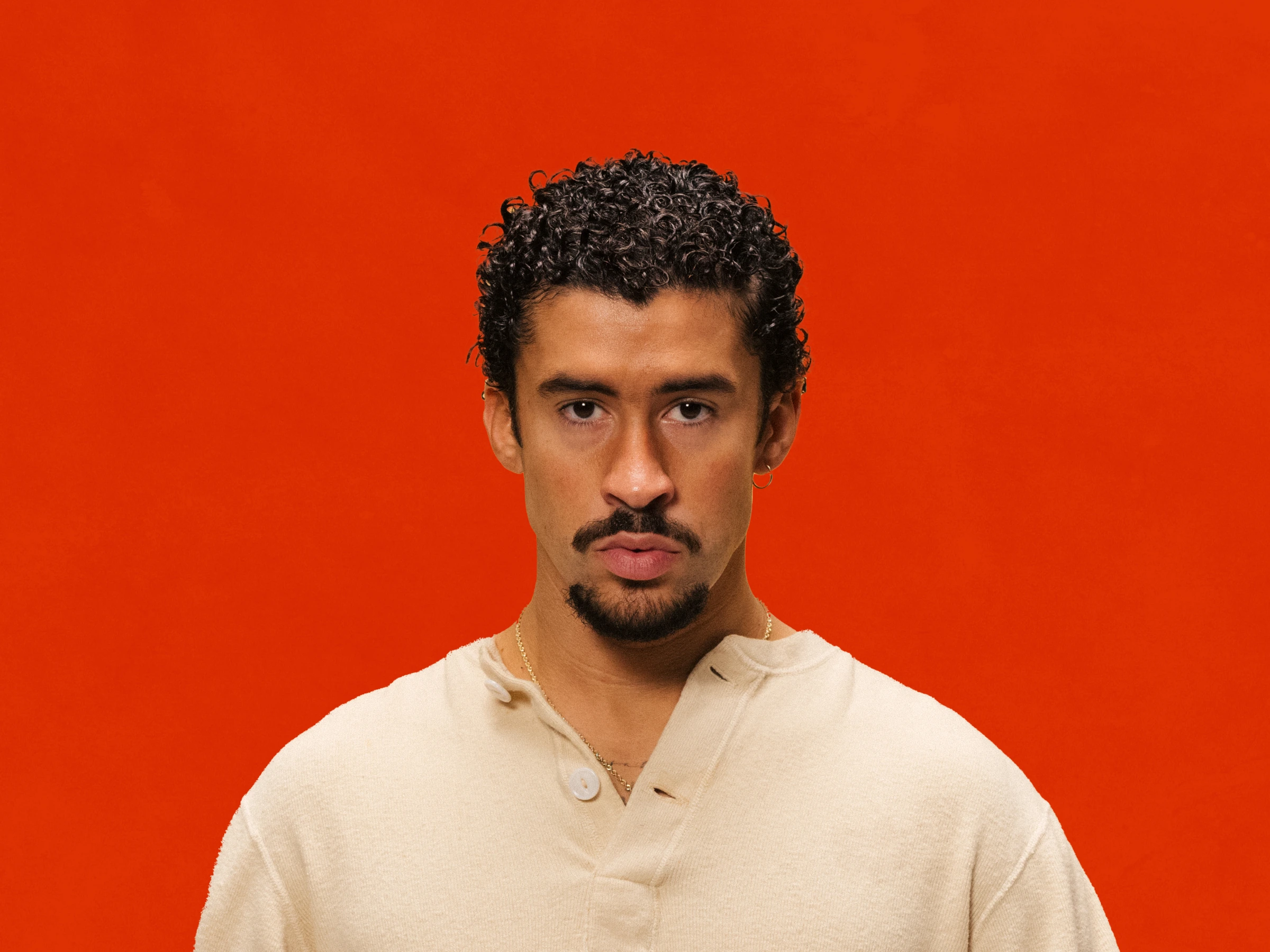 Bad Bunny, who has curly hair and a goatee, wearing a beige shirt, stands against an orange background.