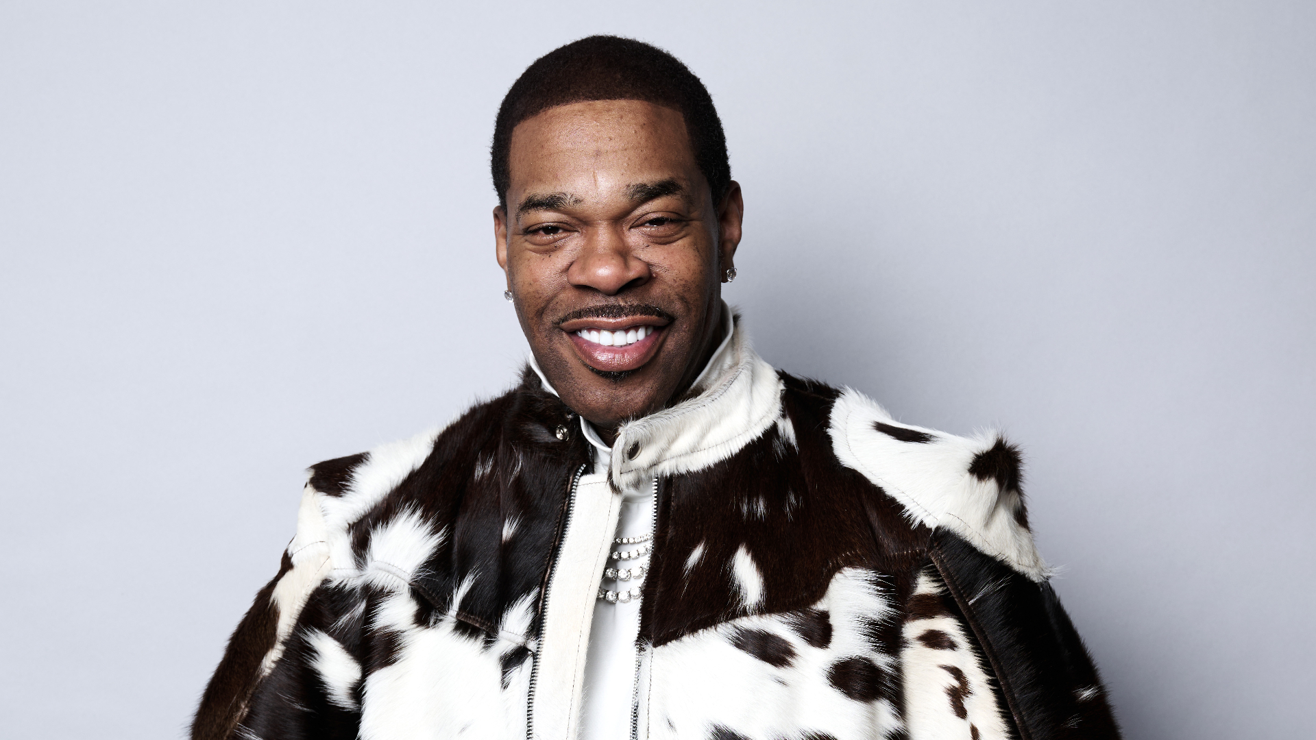 Busta Rhymes smiling in a stylish black and white fur coat against a plain background.