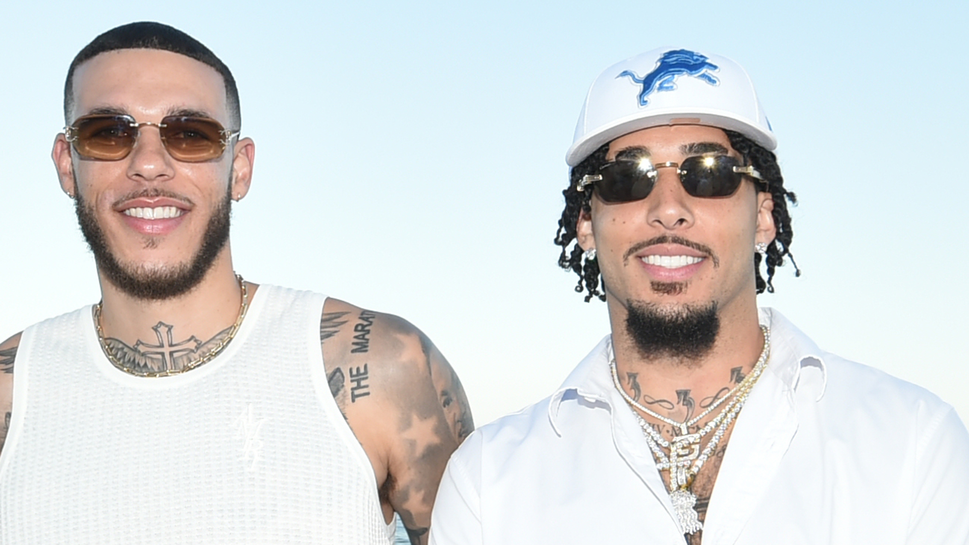Lonzo and LiAngelo Ball smiling, wearing sunglasses. Lonzo in a white tank top with tattoos, LiAngelo in a white shirt and Detroit Lions cap.
