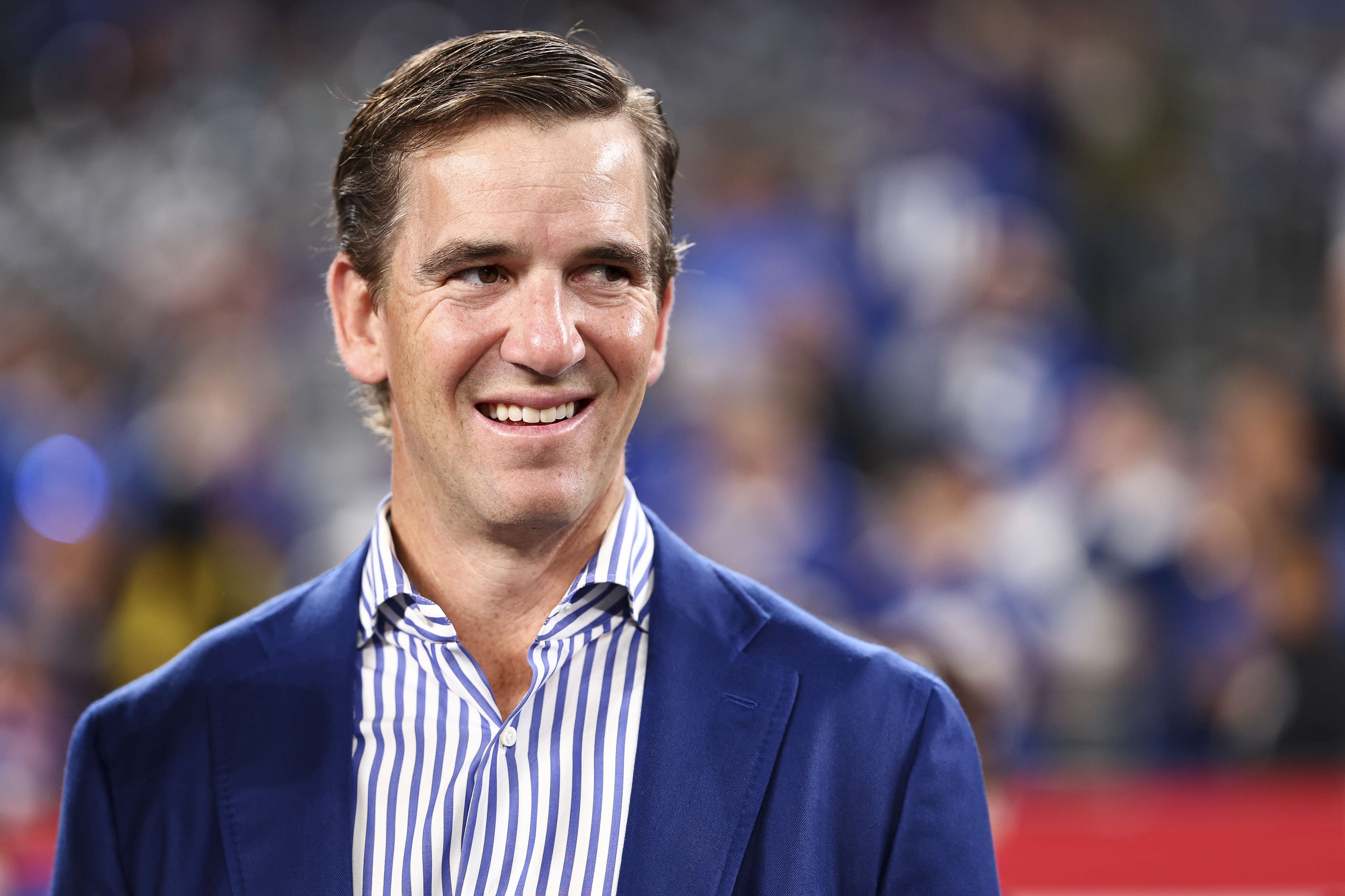 Eli Manning, former NFL quarterback, wearing a blue blazer and striped shirt, smiles at an event.