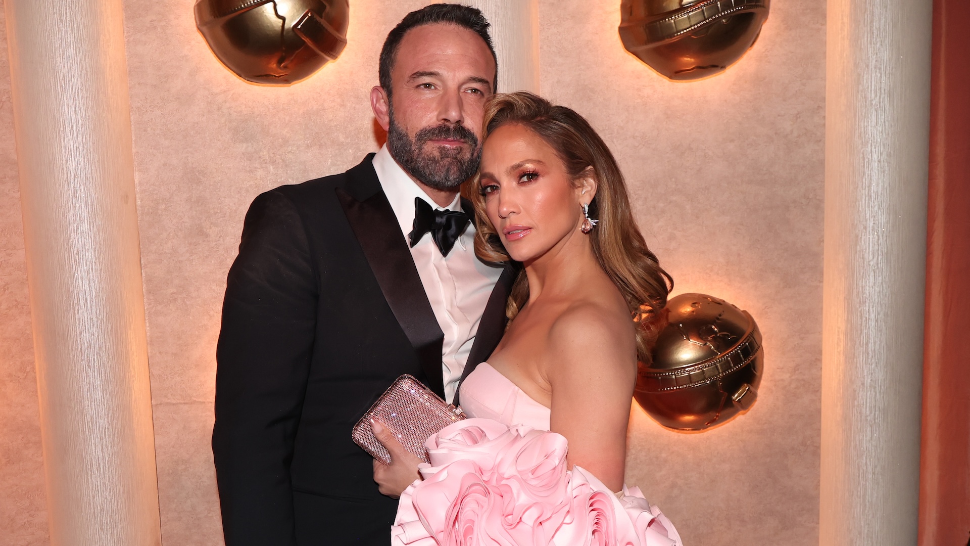 Ben Affleck in a tuxedo and Jennifer Lopez in a pink dress pose together at an event with golden decor in the background.