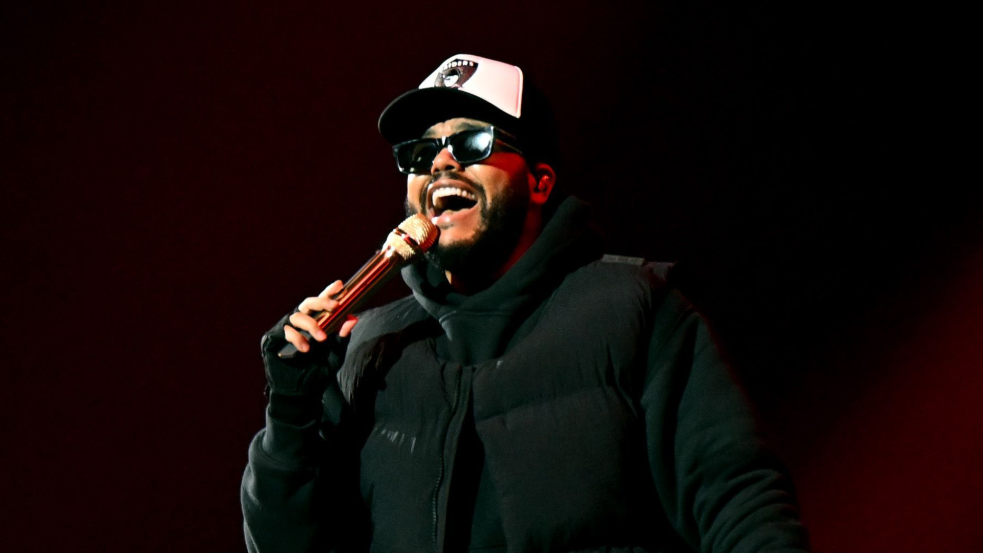 A man wearing sunglasses and a cap performs on stage, holding a microphone. The background is dark, and he is dressed in a black outfit.