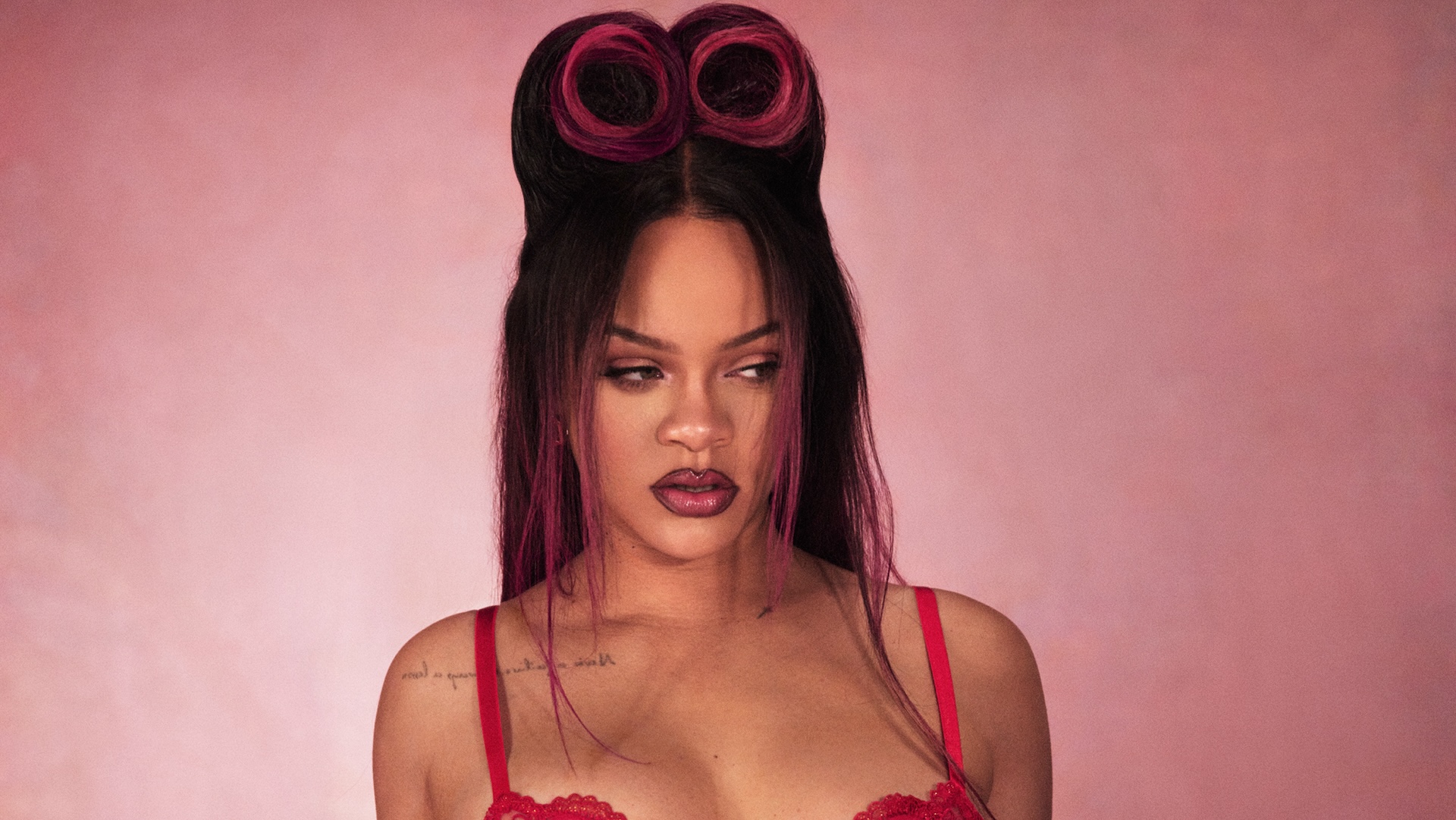 Rihanna in a red outfit with styled hair and a pink background.
