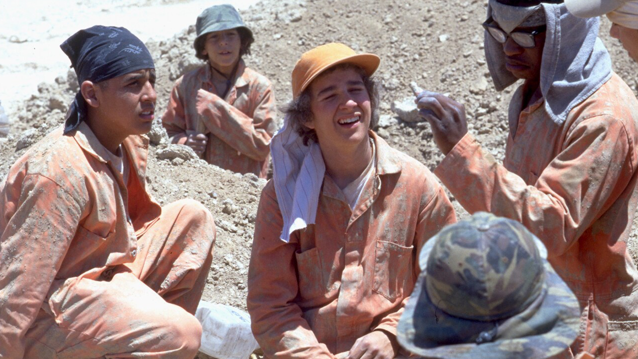 Still from the 2003 film "Holes." A group of young men, including Shia LeBeouf at center, in orange jumpsuits sit in a dusty area, appearing to be engaged in conversation or a break.