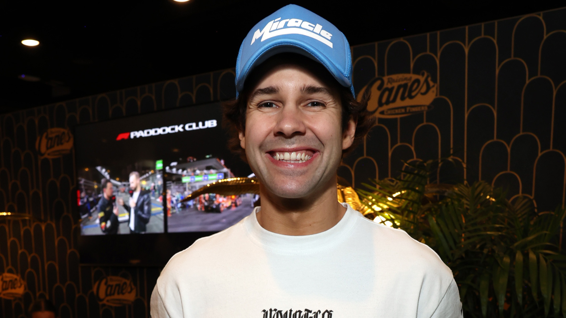 David Dobrik smiling, wearing a blue cap and a white shirt, standing indoors with a screen in the background.