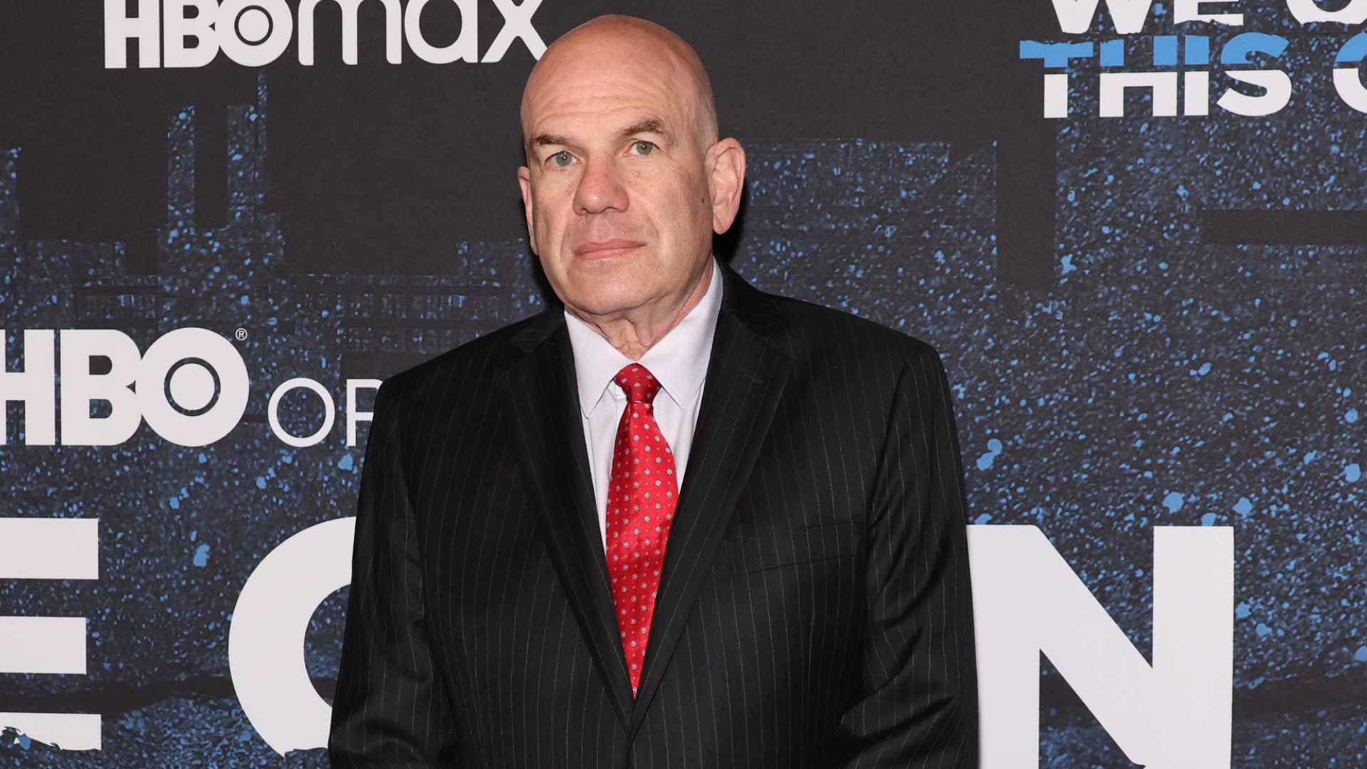 David Simon in a suit with a red tie stands in front of an HBO Max backdrop at an event.
