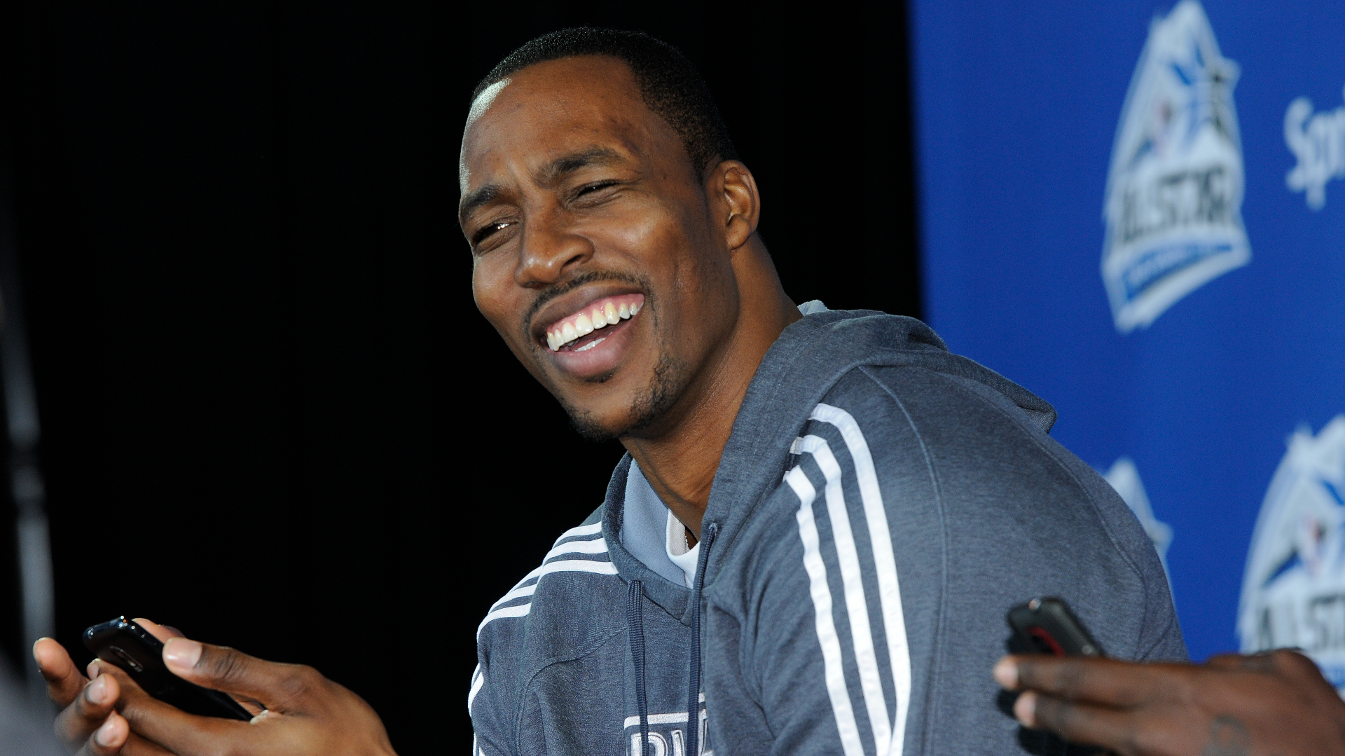 Dwight Howard smiling in a press conference, wearing a gray hoodie, with a phone in his hand.