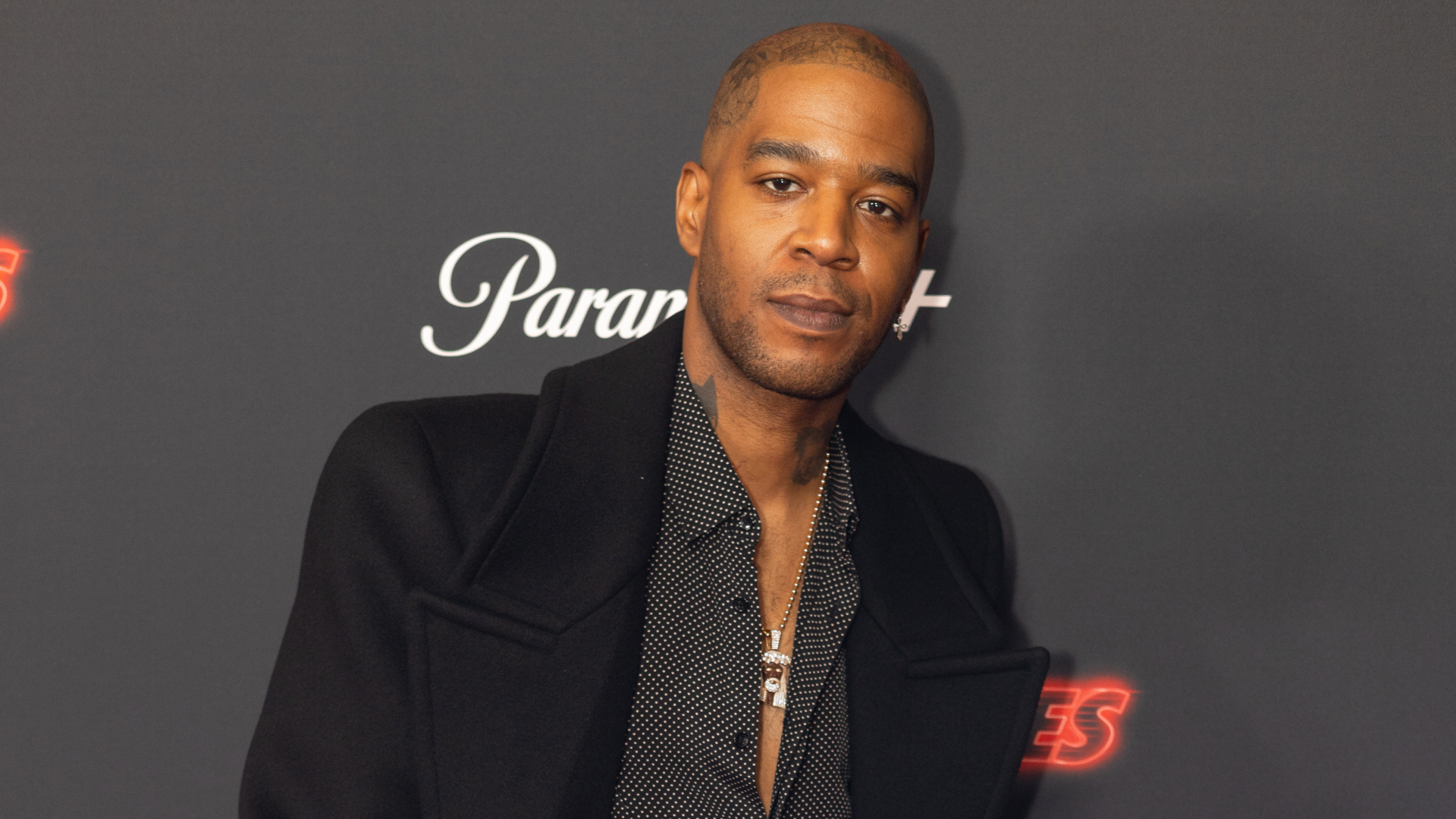 Kid Cudi at an event, wearing a black coat over a polka dot shirt, standing against a dark background with logos.