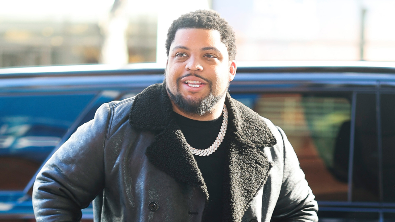 O'Shea Jackson Jr. wearing a black shearling coat and a chain necklace stands outdoors, smiling, with a car in the background.