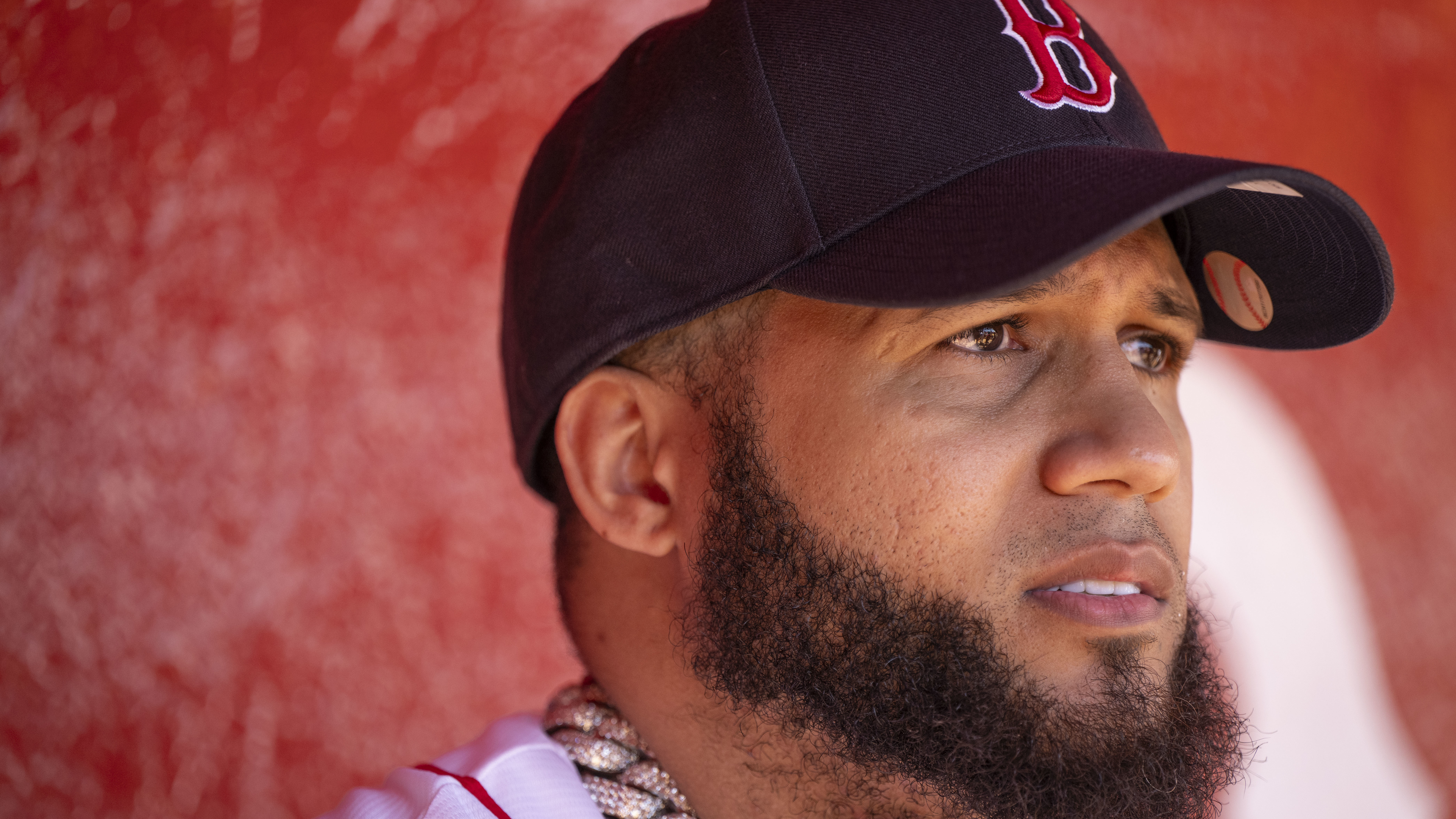 El Alfa with a thick beard wears a Boston Red Sox cap and a white jersey, standing against a red background.