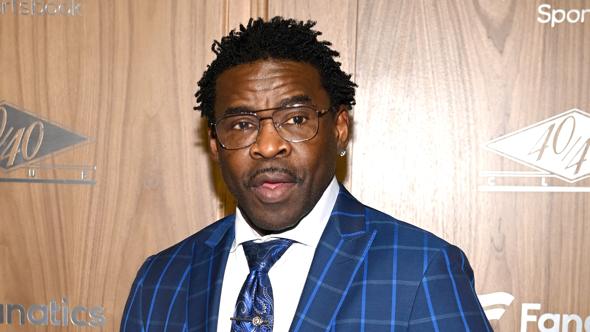 Michael Irvin wearing a blue checkered suit and glasses, standing against a wooden backdrop.