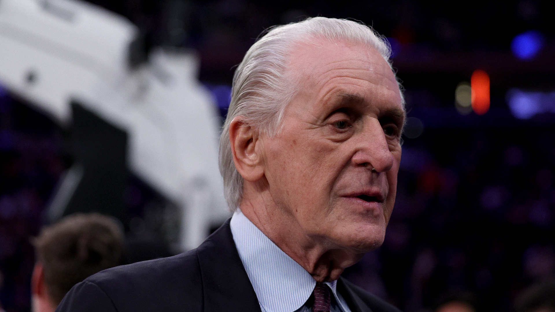 Pat Riley in a suit, standing in a dimly lit arena.