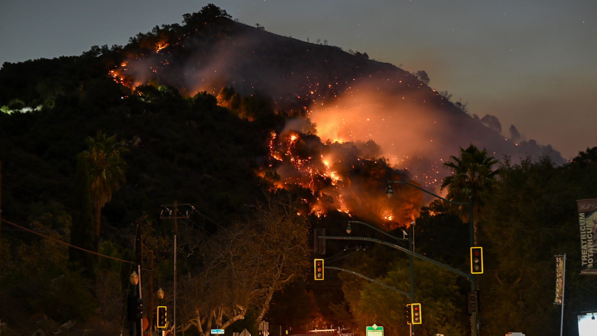 A hillside is engulfed in flames during a wildfire at night, with smoke billowing into the sky. Traffic lights are visible in the foreground.