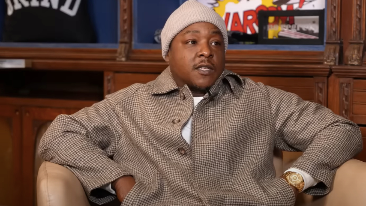 Jadakiss in a houndstooth coat and beanie sits in an armchair, with a colorful background.