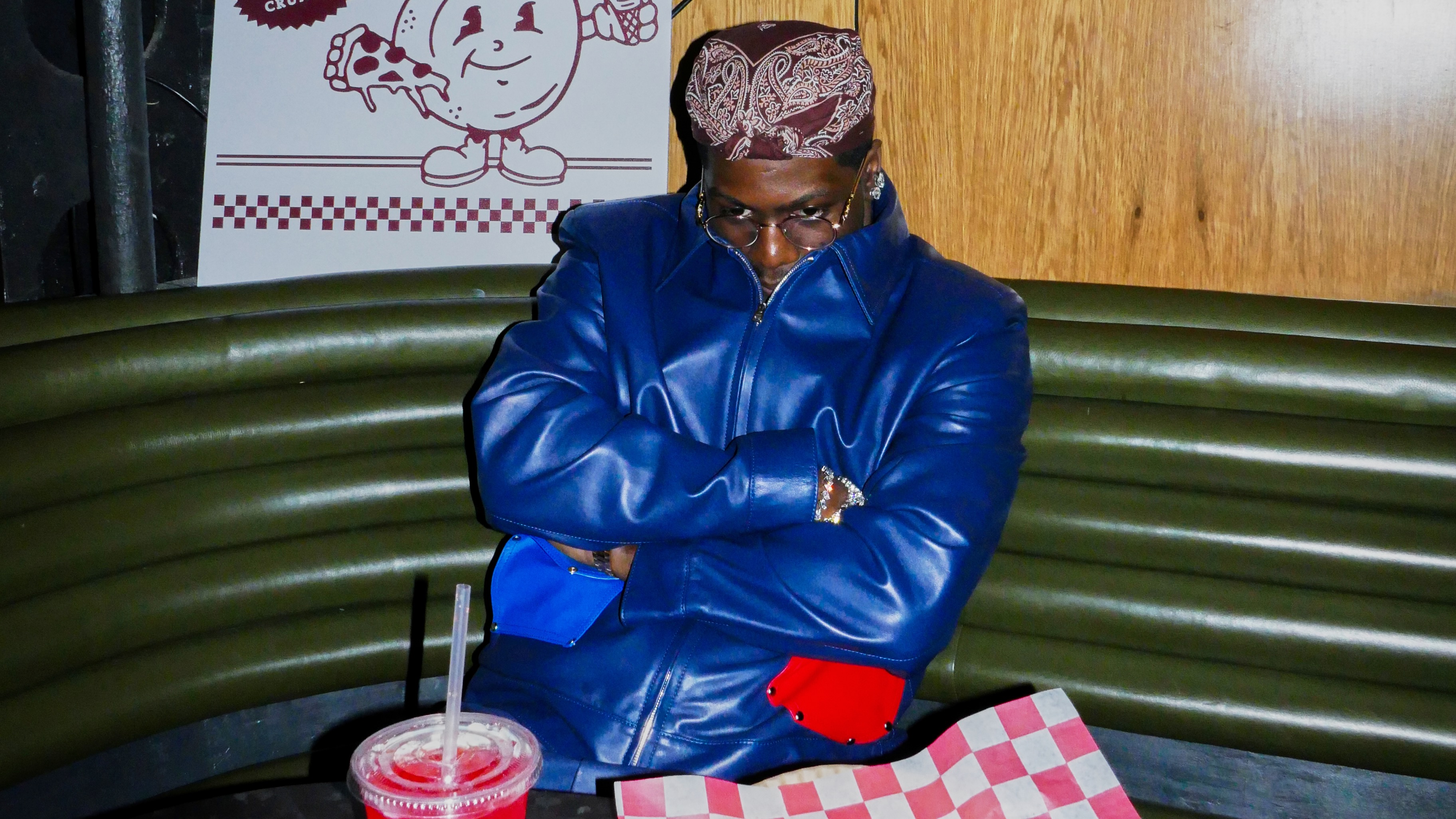 A person in a blue jacket and bandana sits with arms crossed in a booth, next to a drink and an empty tray with a checkered paper.